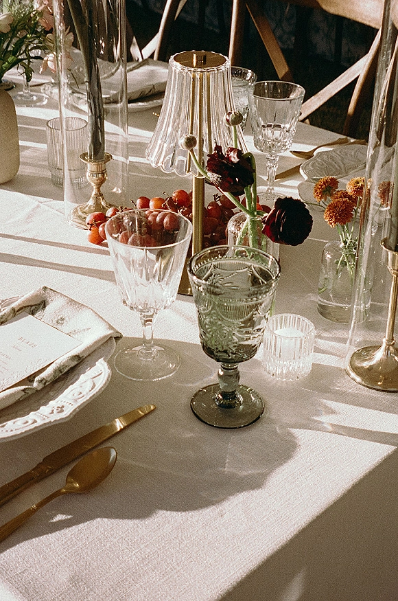 Reception tablescape with wedding table setting on a white tablecloth, brass flatware, scalloped plates, taper candles, bud vases, grapes in sunlit shadows