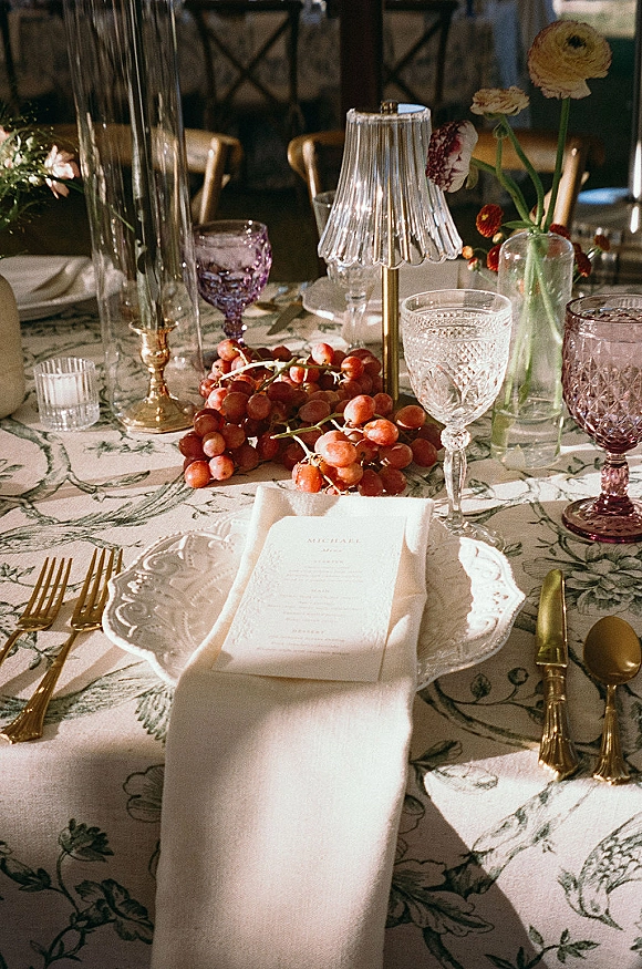 Reception tablescape with wedding place setting on patterned tablecloth, scalloped plates, menu card, gold flatware, crystal goblets, taper candles.