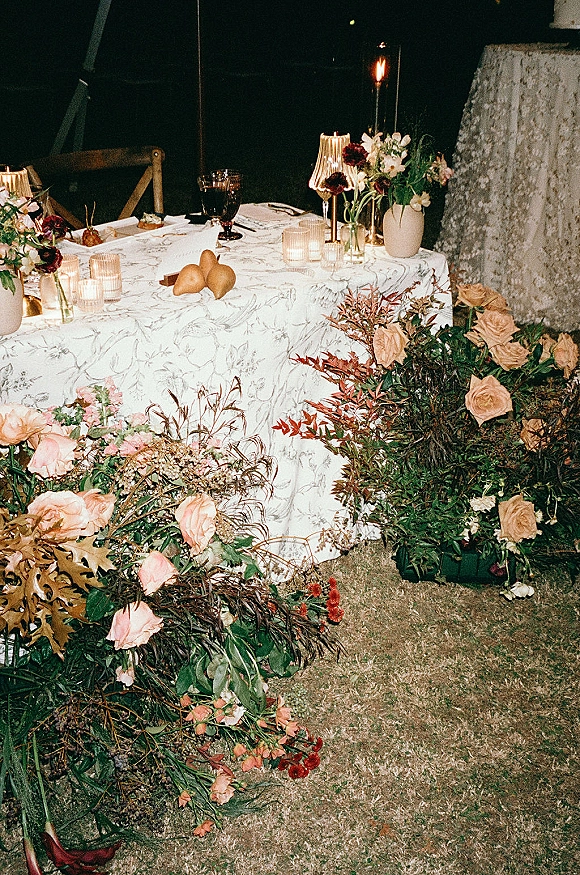Reception tablescape with candlelit reception table details, patterned tablecloth, taper candles, roses and pears on an outdoor lawn at night