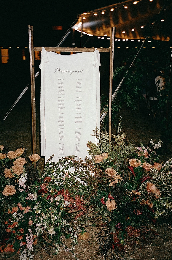 Seating chart display with wedding seating chart on fabric in a wooden frame, with roses and greenery under tent string lights at night