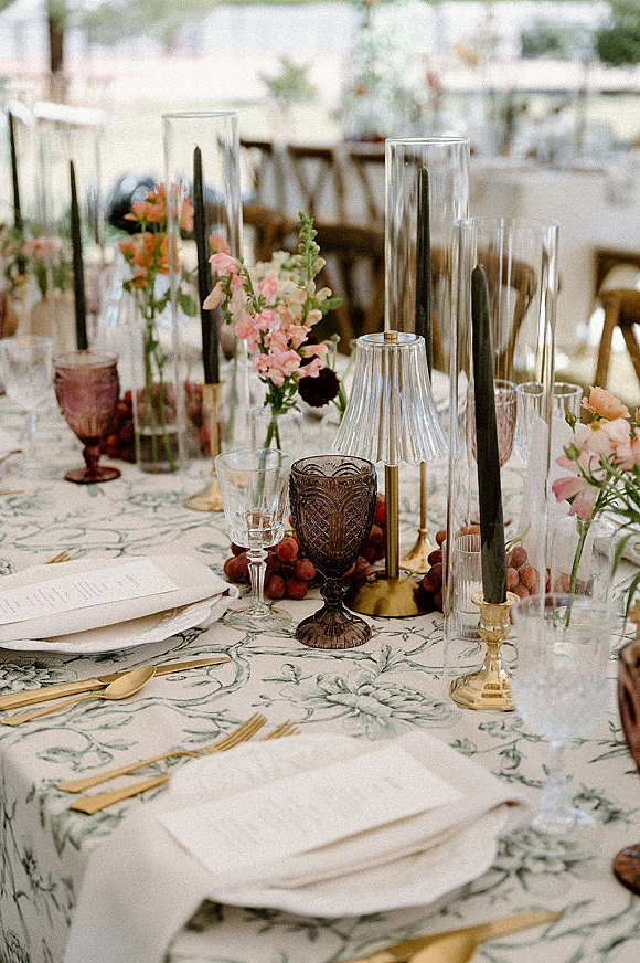 Reception tablescape with wedding table setting on patterned tablecloth, bud vase florals, black taper candles, grapes, and tented seating behind