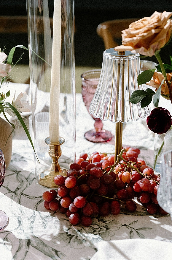 Reception tablescape with wedding table decor featuring roses, greenery, grapes, goblets and taper candles in brass holders on a patterned cloth in dim interior