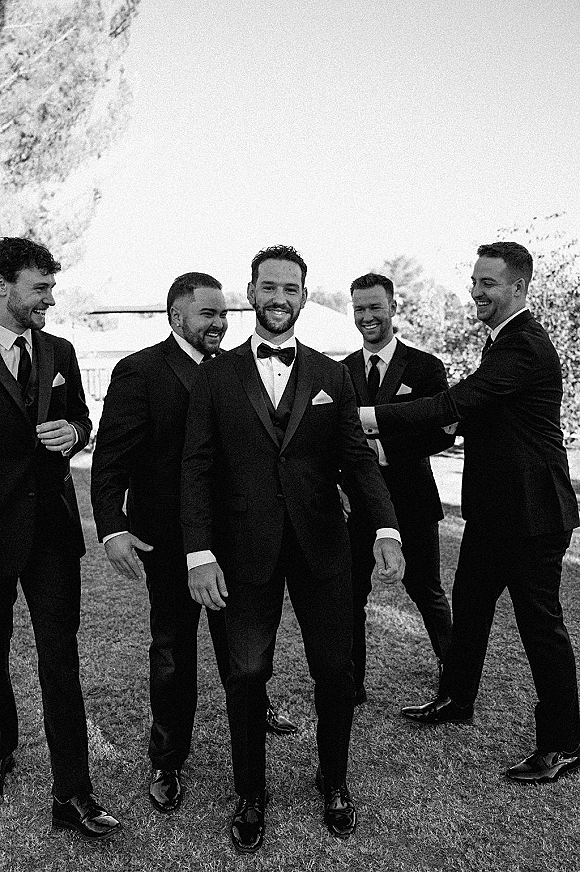 Groom with groomsmen laughing in black tuxedos and bow ties, boutonniere and pocket squares, standing on a lawn with trees and bright sky