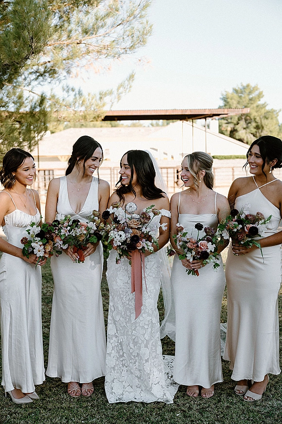 Bridesmaid group portrait with bride with bridesmaids in ivory dresses holding bouquets with floral ribbon on a grassy lawn by trees and fence