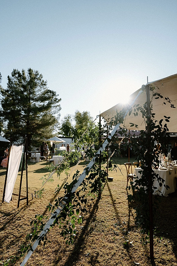 Outdoor wedding reception under a sailcloth wedding tent with greenery garland and string lights over tables on a sunny lawn