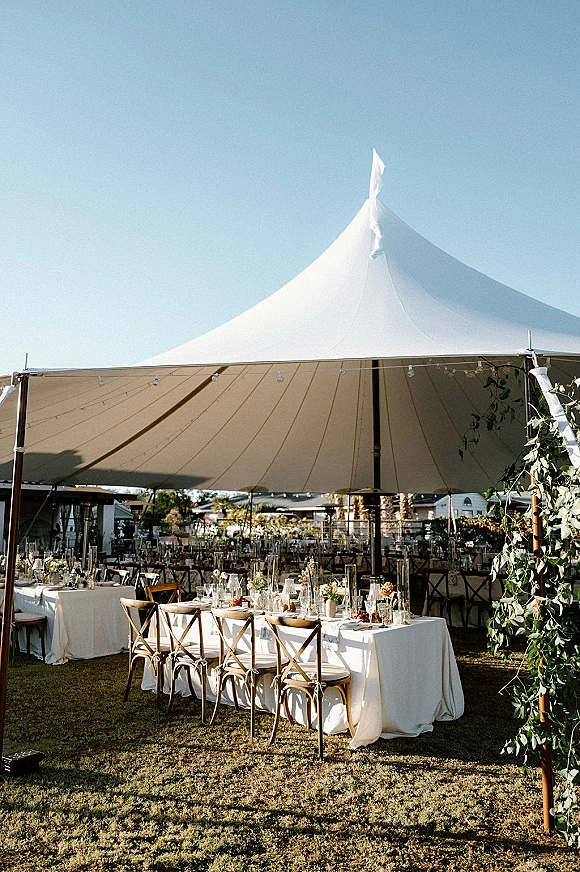 Tent reception decor with sailcloth wedding tent, string lights, and long banquet tables set with white linens, florals, and taper candles on a lawn