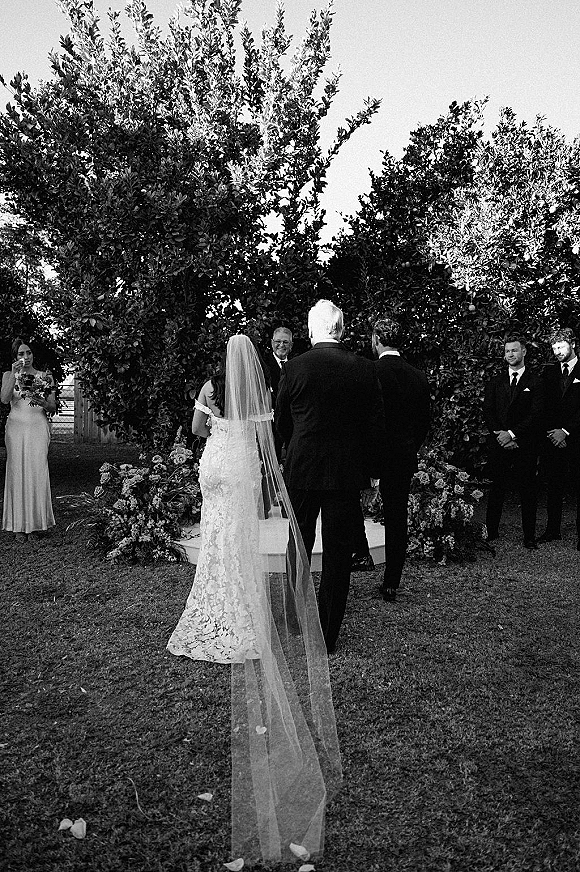 Ceremony moment as bride walks down the aisle in a long veil and lace dress, with bridesmaids, groomsmen, and floral-lined garden lawn backdrop