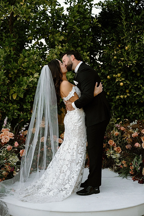Wedding kiss portrait of bride and groom kiss, her lace dress and long veil flowing by floral ground arrangement in a citrus grove