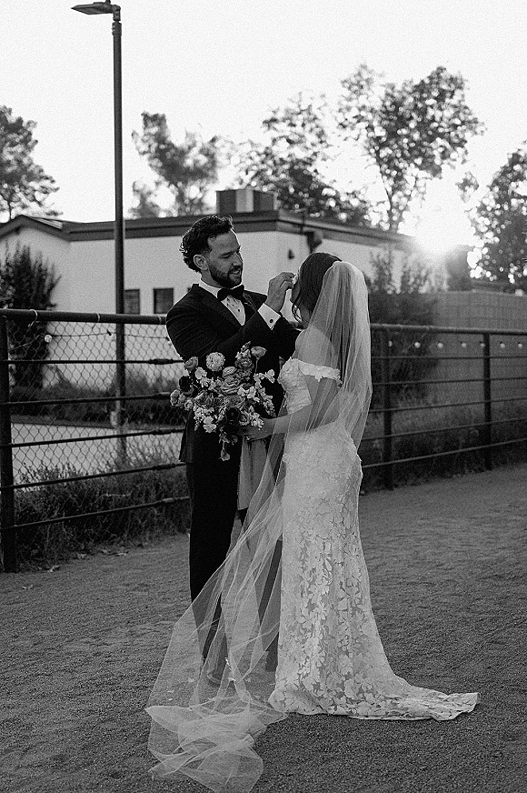 Couple portrait of bride holding a bouquet as groom adjusts her veil, cathedral train glowing in sun flare by an urban fence path