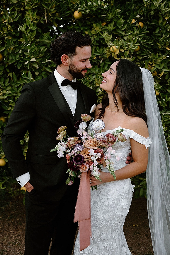 Couple portrait of bride and groom holding a bouquet with blush ribbon, gazing at each other beneath orange trees in a citrus grove