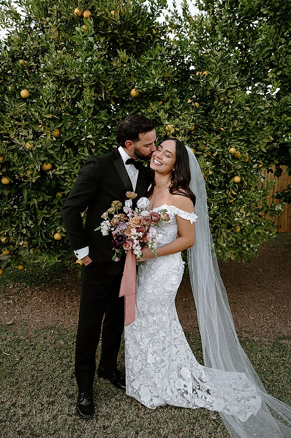 Couple portrait of bride and groom kiss, her lace dress and long veil flowing as she holds a bouquet in a citrus garden backdrop