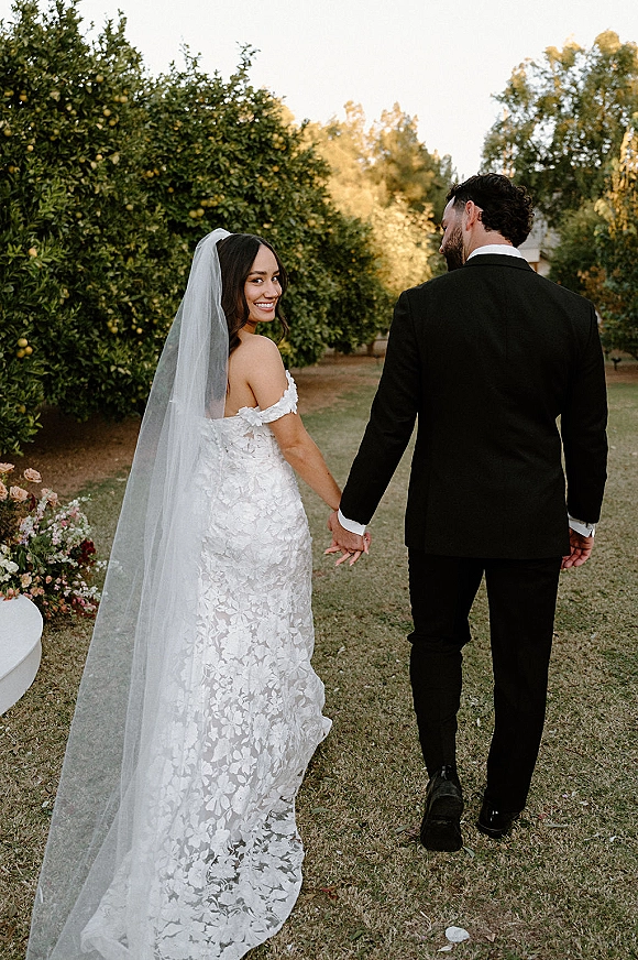 Couple portrait of bride and groom holding hands, walking away in an orchard at sunset, her long cathedral veil flowing behind