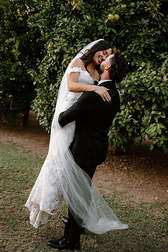 Wedding couple portrait of groom lifting bride in lace gown with long veil, embracing on a dirt path beside green orchard foliage