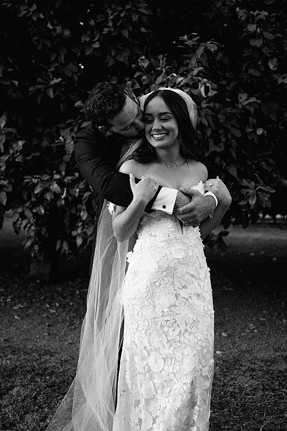 Couple portrait in a black and white wedding portrait, groom kissing bride’s forehead as she smiles in a cathedral veil amid garden trees
