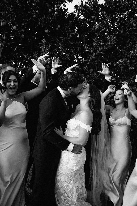 Wedding kiss as the bride in a lace off-the-shoulder gown and veil leans into the groom in a tux, wedding party cheering amid garden trees