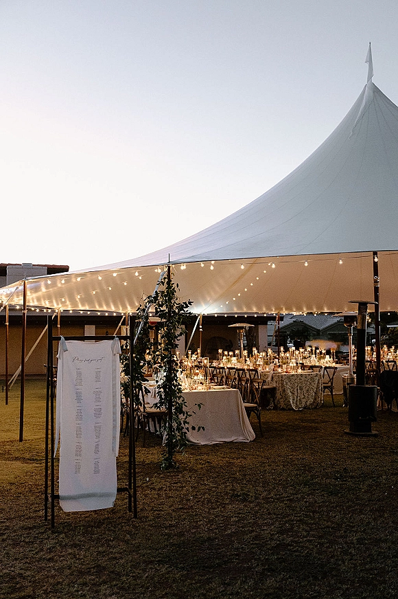 Wedding reception tent with sailcloth canopy, string lights, banquet tables, candles, and greenery on a lawn under evening sky