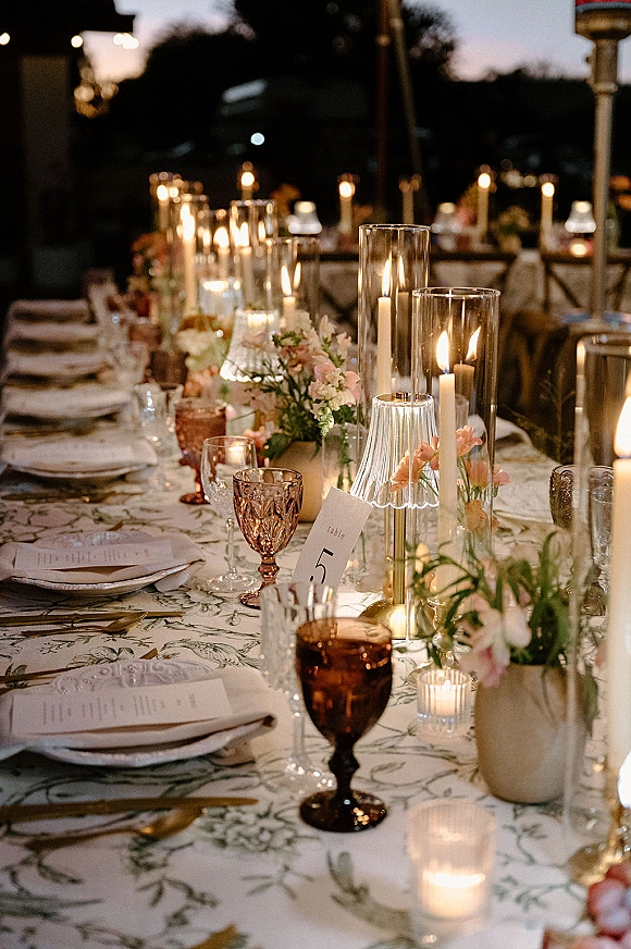Reception tablescape with a candlelit wedding table of taper candles, hurricane vases, florals, and amber goblets under bistro string lights outdoors at night