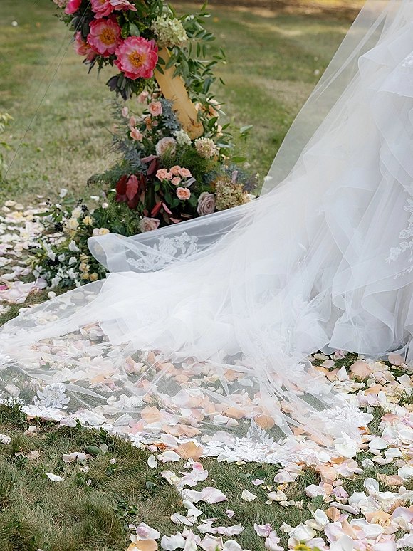 Wedding dress train with lace wedding train detail, tulle skirt and veil draped on grass beside blush rose petal aisle and floral arch greenery