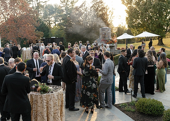 Cocktail hour wedding cocktail hour guests mingling by high-top tables with patterned linens, signature drinks, and a smoking stone fireplace outdoors