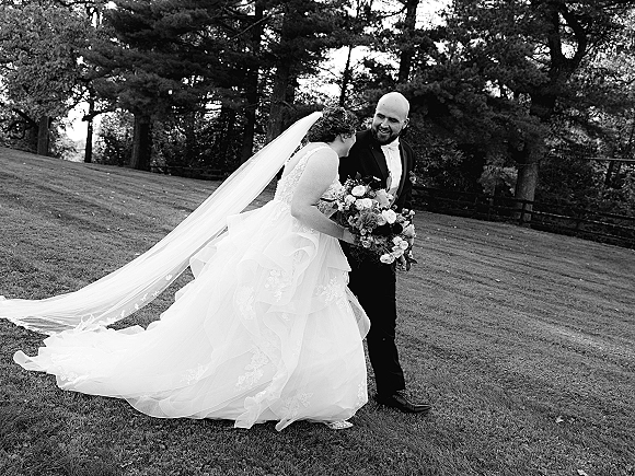 Couple portrait in a black and white wedding portrait, bride laughing and leaning on groom as her long veil trails on a lawn with trees