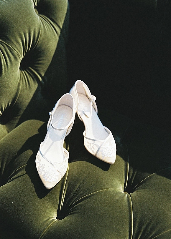 Wedding shoes bridal heels with ankle straps and lace appliqué resting on a green velvet tufted chair in soft sunlight shadows