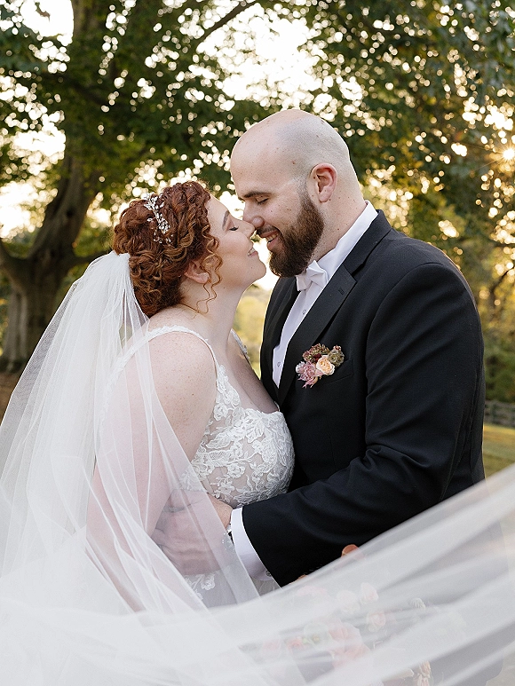 Wedding kiss portrait of bride and groom kissing as her veil blows, lace dress and floral hairpiece lit by sunlight among trees