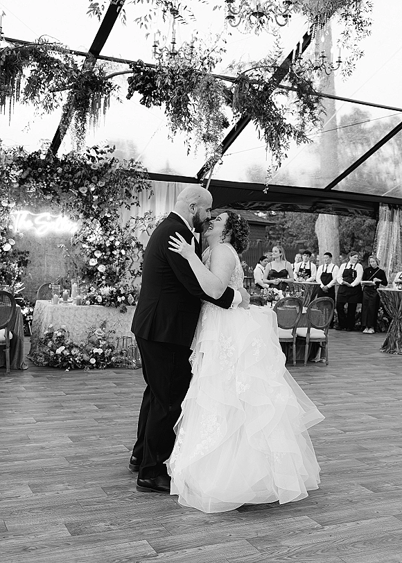 First dance as the wedding couple dancing on a wooden floor under a clear top tent, chandelier and hanging greenery, guests watching