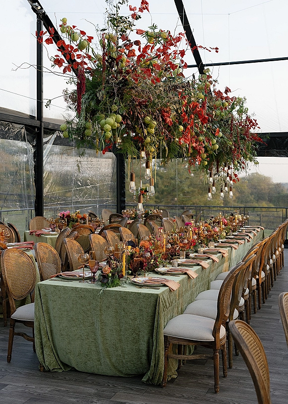 Reception tablescape with green wedding table linens on a long banquet table, taper candles and florals under Edison bulbs in a glass greenhouse