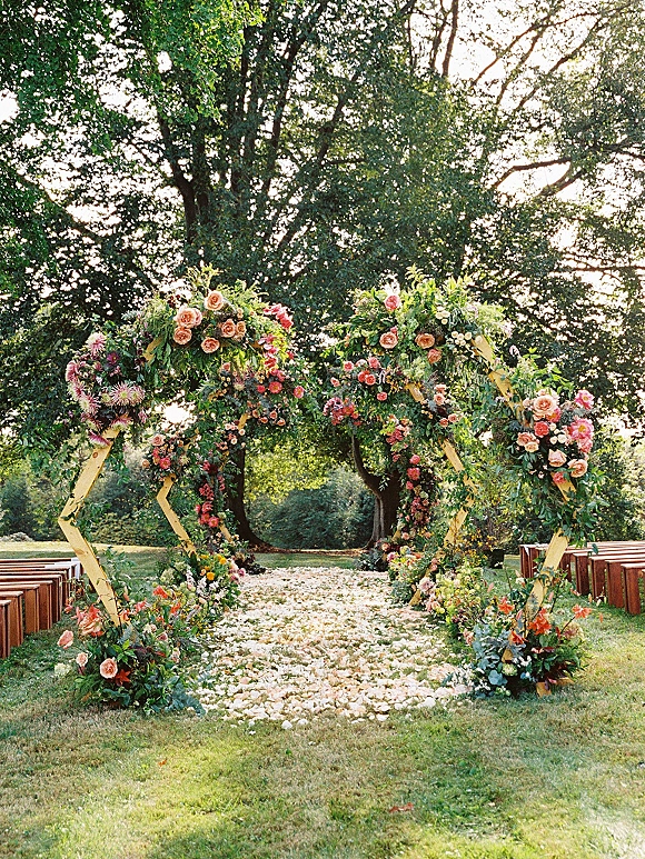 Ceremony altar with an outdoor wedding ceremony setup featuring hexagon wedding arch, garden roses and greenery on a lawn under trees