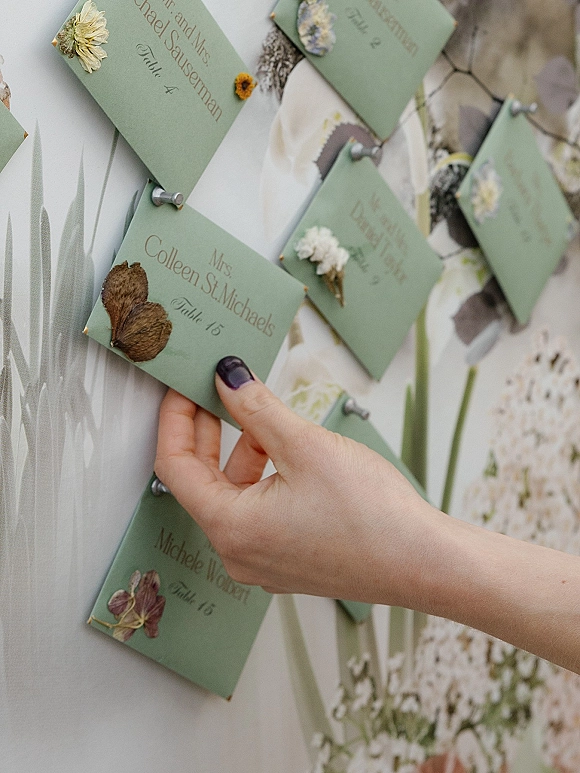 Seating chart display with wedding escort cards, green escort cards pinned with pressed flowers and dried leaves on a botanical board by a light wall