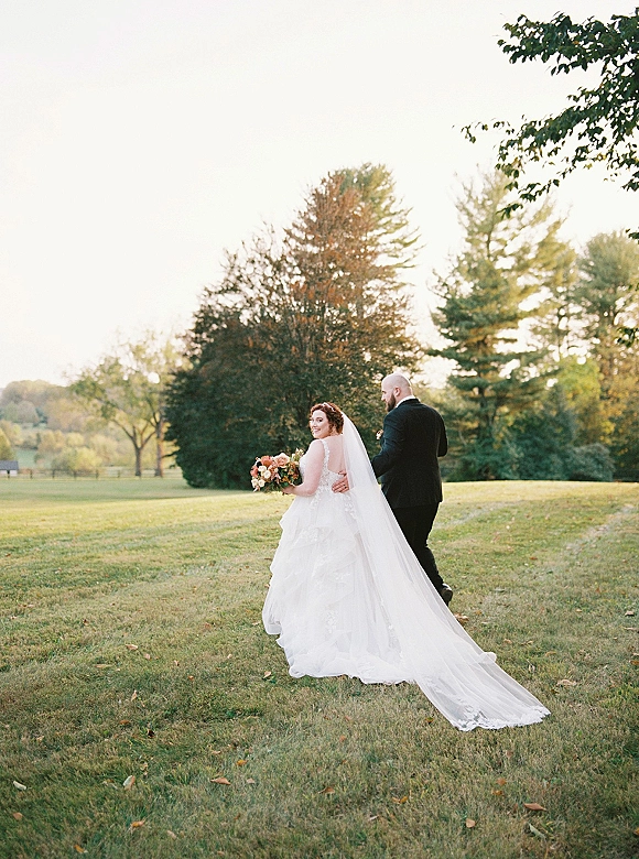 Couple portrait of bride and groom walking away on a park lawn, bride in long veil holding a bouquet as he holds her waist