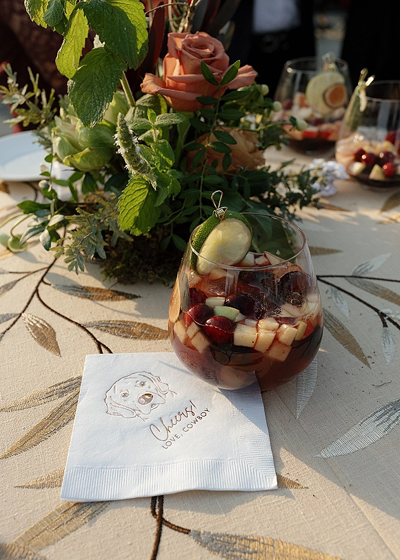 Reception tablescape with wedding table centerpiece of roses, moss and greenery, plus fruit cocktails with lime garnishes on leaf-print linens outdoors