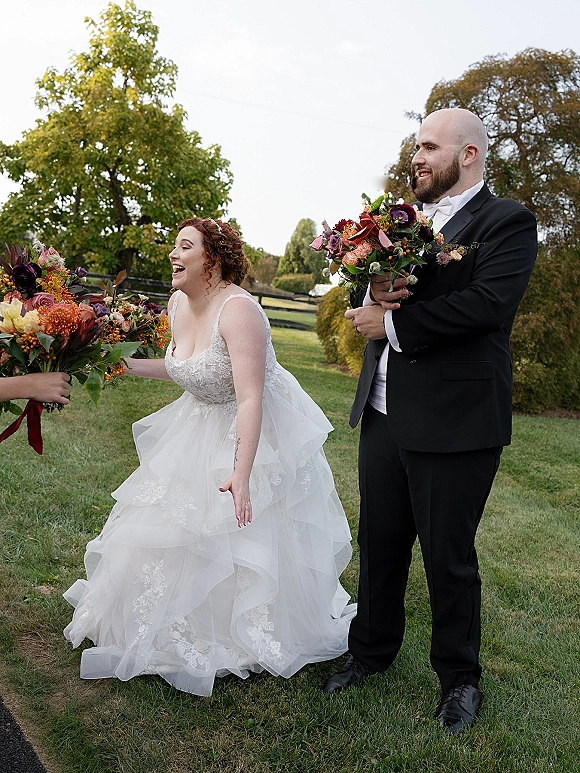 Wedding party photo of a laughing bride and groom with bouquets as a bridesmaid hands flowers on a grassy lawn with trees and fence
