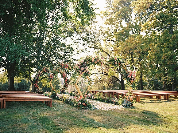 Ceremony setup for an outdoor wedding ceremony with triple hexagon arches, lush florals and petals lining a garden aisle under trees