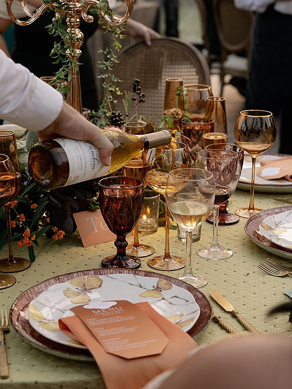 Reception tablescape wedding place setting with amber glass goblets, gold flatware, taper candles, candelabra, and greenery garland as guests sit behind