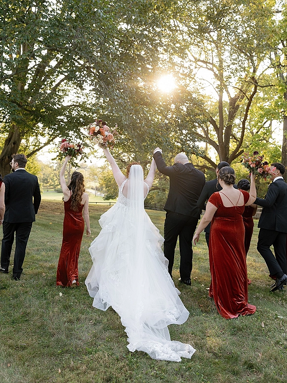 Wedding party portrait of bride and groom walking away hand in hand, bridal party raising bouquets on a sunlit park lawn among trees