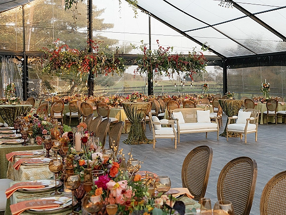Reception tablescape under a clear tent reception with long green-clothed banquet tables, orange napkins, candlelit florals, and string lights overhead