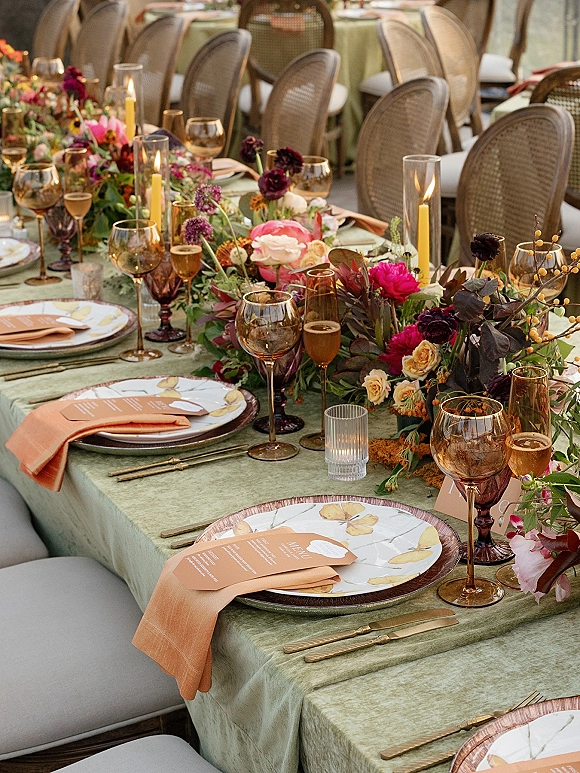 Reception tablescape with wedding table centerpiece garland, taper candles in hurricane vases, patterned plates, menus and place cards on wine glasses, cane chairs behind