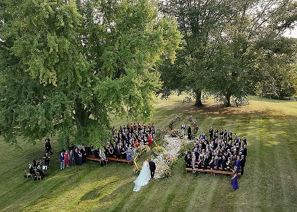 Outdoor wedding ceremony with a petal aisle leading to a floral ceremony arch, wooden benches in a semicircle on a tree-lined lawn
