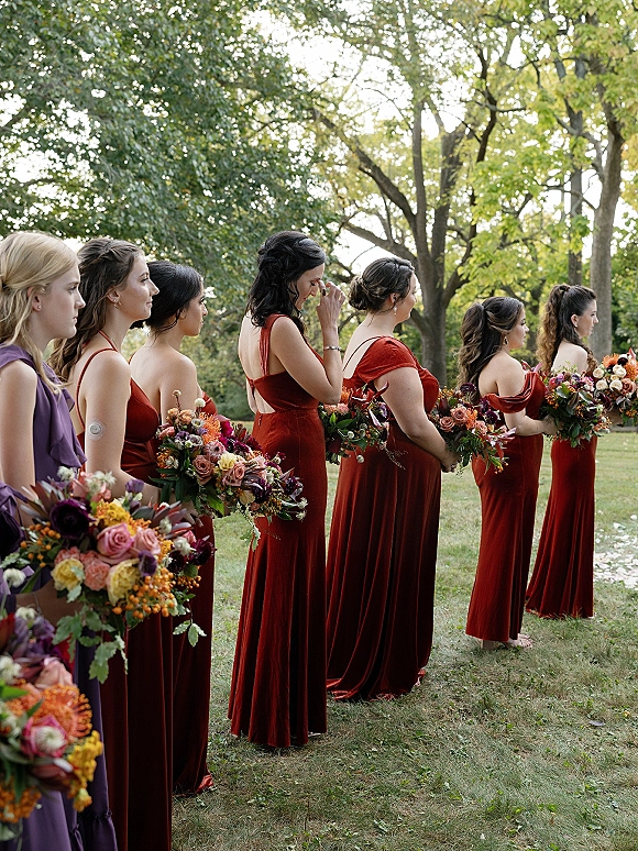 Bridesmaid lineup in rust dresses holding bouquets, shown in side profile on a grassy park lawn with trees, earrings visible