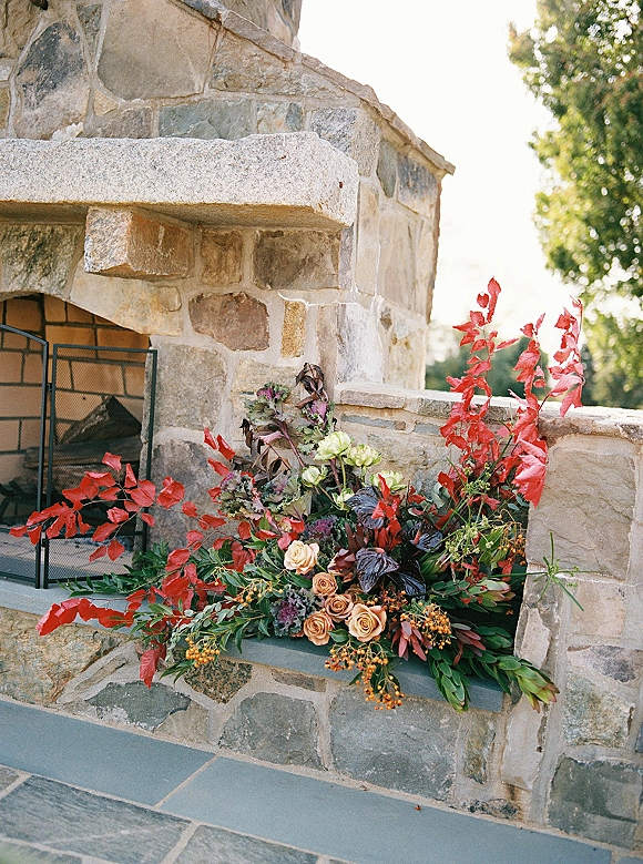 Wedding floral installation with roses and greenery cascading over a stone fireplace mantel, with red leaves and orange berries outdoors