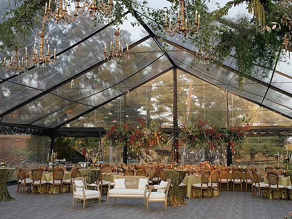 Reception tablescape under a clear tent reception with chandeliers, hanging greenery, long banquet tables, taper candles, and dusk trees beyond
