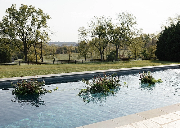 Floating florals drift on a pool with greenery accents, set on a stone deck with rolling hills, split rail fence, and trees beyond