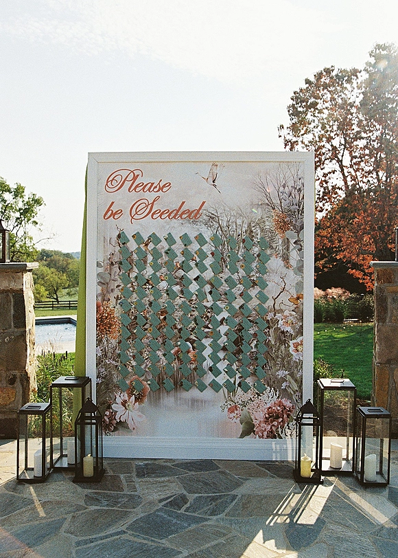 Wedding seating chart with escort card display on a floral illustrated backdrop, accented by lanterns on a stone patio with columns and lawn