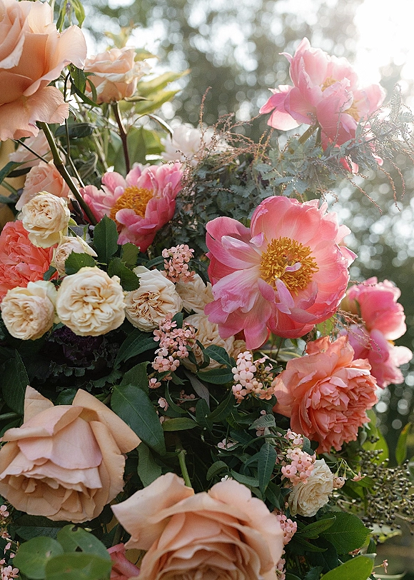 Wedding floral arrangement of peony wedding flowers with roses, spray roses, and greenery, lit by sunlight with trees and sky bokeh behind