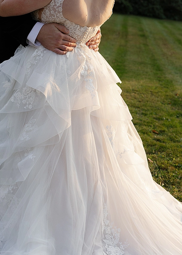 Wedding dress close-up showing lace wedding dress detail with floral appliqués and tulle ruffles as groom’s hands rest at her waist outdoors