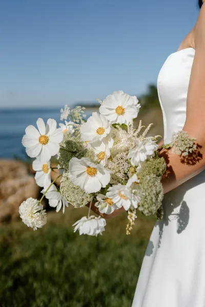 Bridal bouquet of white cosmos bouquet and wildflowers with greenery, held against an ocean coastline under a blue sky in grass