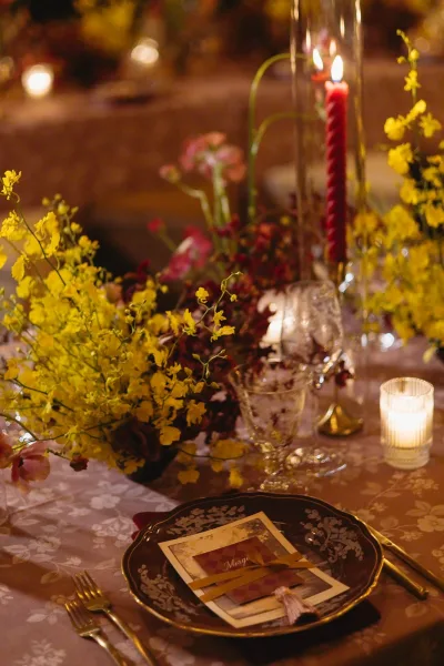 Reception tablescape with yellow floral centerpiece and a red taper candle glowing against a patterned tablecloth at a candlelit dinner.