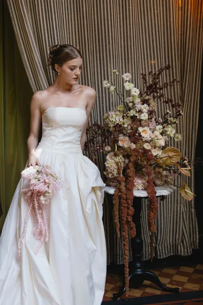Bridal portrait of a bride holding bouquet in a strapless gown, set against striped drapery and a checkered tile floor.