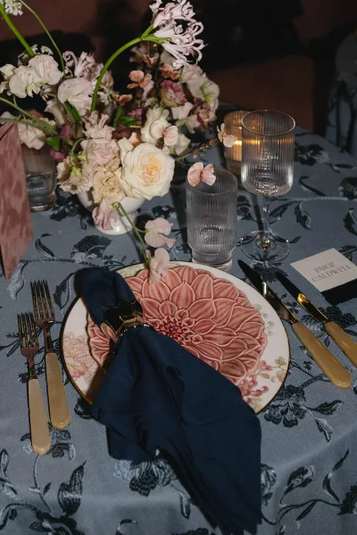 Wedding place setting in a blue and blush tablescape, with a gold napkin ring at the plate against a dark backdrop.